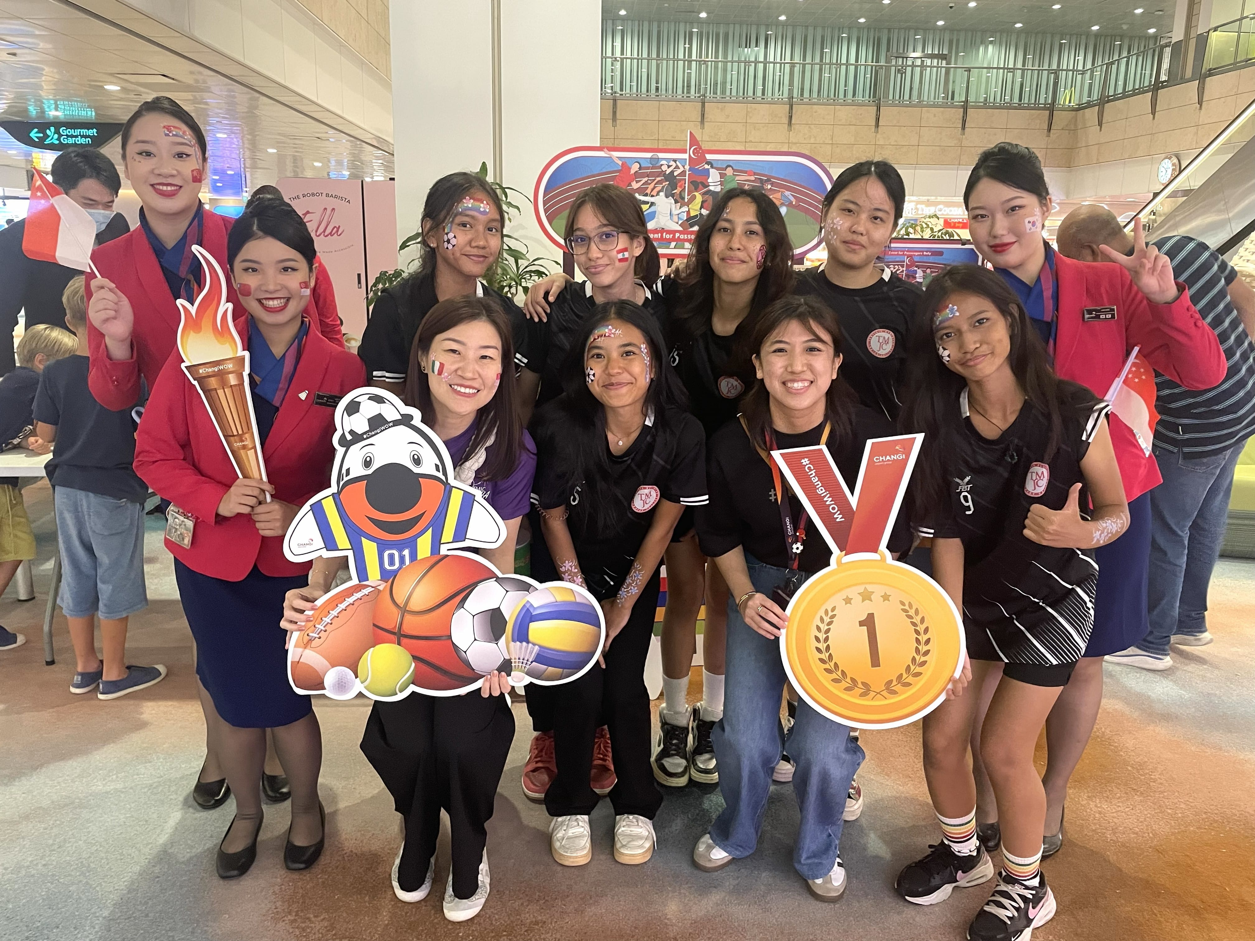 A group of students in black TMJC sports jerseys pose with airline staff in red uniforms at what appears to be Changi Airport. Several participants have flag face paint and hold #ChangiWOW props including a gold medal and sports mascot cutout.